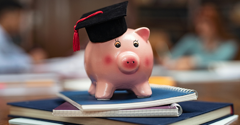 Pink ceramic pig figurine with a black graduate cap on top of notebooks indicating higher education and study loans